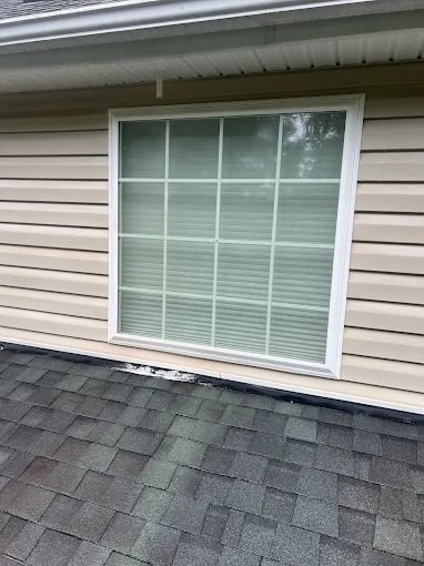 White window with blinds on beige siding and gray shingled roof